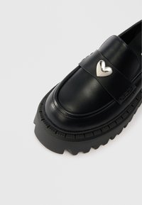 Black leather loafers with a chunky textured sole and a silver heart-shaped accent on the strap. Smooth finish and raised stitching details.