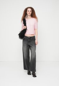 Γυναίκα με σγουρά μαλλιά που φοράει ροζ ζακέτα με κοντά μανίκια, ξεθωριασμένο μαύρο τζιν με φαρδιά πόδια, μαύρες μπότες και κρατάει μια μαύρη χνουδωτή τσάντα.