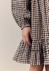 Kárované šaty v béžovej, hnedej a modrej farbe s nafúknutými rukávmi a volánovou lemovkou. Detail ruky spočívajúcej na šatách.