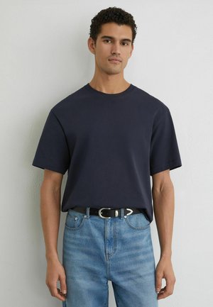 Model masculin cu păr creț purtând un tricou bleumarin băgat în blugi bleu deschis cu o curea neagră, stând în fața unui perete alb simplu.