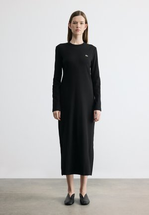 Femme debout, portant une longue robe noire à manches longues et des chaussures noires sans lacets, sur un fond blanc uni.