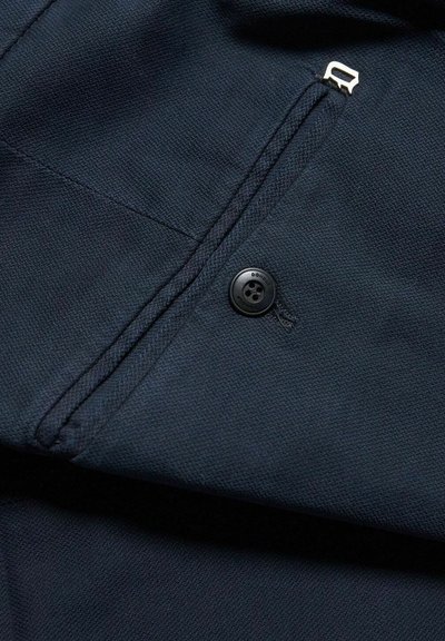 Tessuto della giacca blu navale con una finitura testurizzata. Presenta una cerniera metallica, un bottone nero e cuciture dettagliate lungo le cuciture.