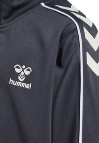 Donkergrijze zip-up jack met witte accenten en logo. Beschikt over een gestructureerde stof en iconisch chevronontwerp op de schouders.