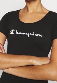 Černé přiléhavé tričko s krátkým rukávem, s bílým kurzívním logem "Champion" na hrudi, vyrobené z měkké bavlny, s kulatým výstřihem.