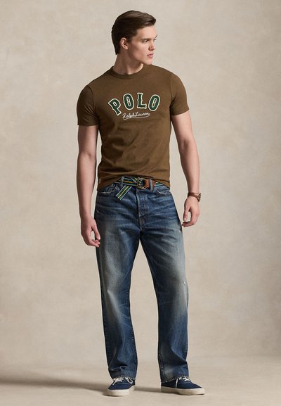 Mladi moški v rjavi majici Polo Ralph Lauren, modrih jeansih s črtastim pasom, mornarsko modrih supergah in z zapestno uro, stoji in gleda desno.