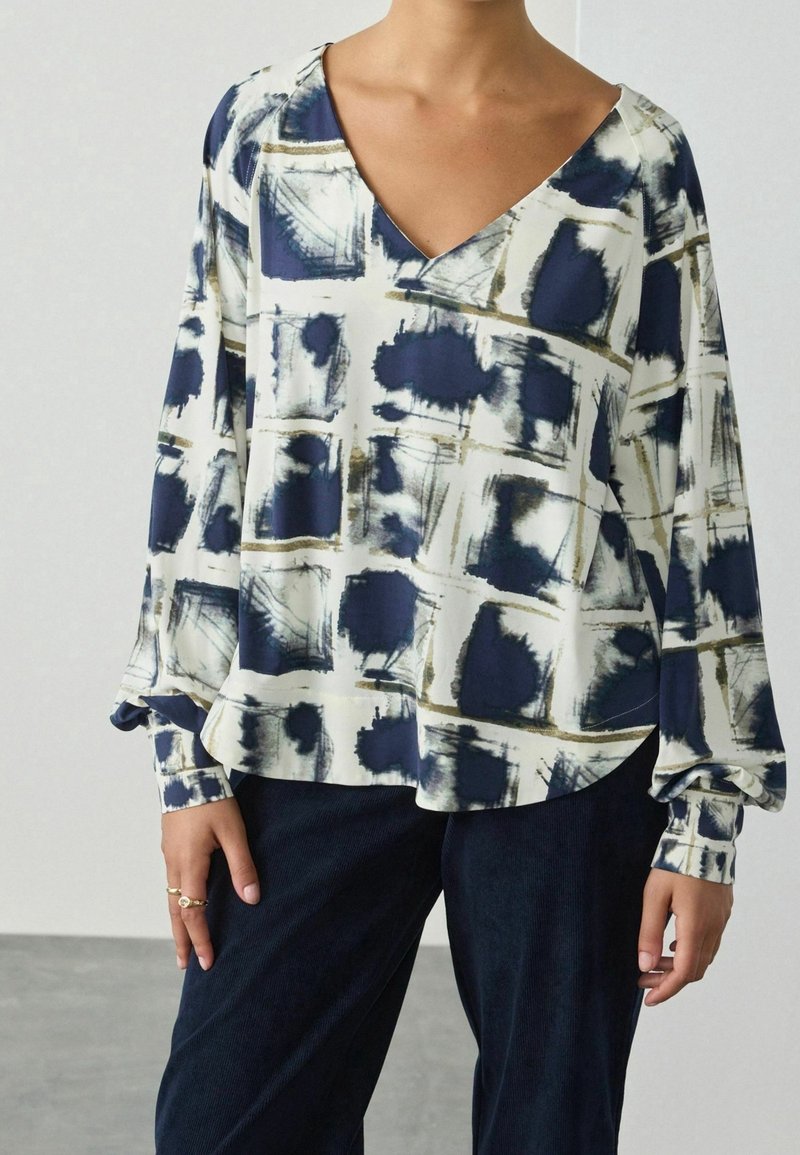 Personne portant un chemisier à manches longues avec un col en V, motifs carrés abstraits bleu marine et blanc, et pantalon foncé.