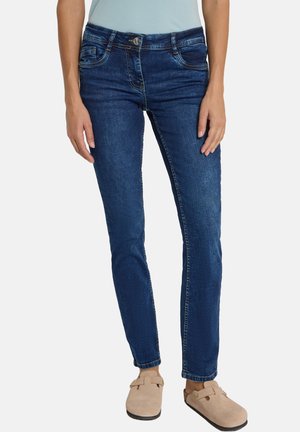 Vrouw draagt donkerblauwe slim-fit jeans en beige instapschoenen, staand tegen een effen lichte achtergrond.