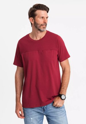 Camiseta roja de manga corta con una sección superior texturizada, cuello redondo y un pequeño bolsillo en el pecho, combinada con jeans azules y un reloj de pulsera.