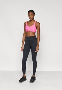 Ροζ αθλητικό σουτιέν με λεπτομέρειες από δίχτυ και μαύρο κολάν με ψηλή μέση. Μοντέλο φορεί μαύρα αθλητικά παπούτσια, ποζάροντας μπροστά σε απλό φόντο.