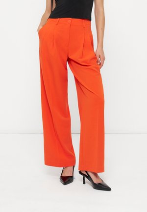 Femme portant un pantalon orange taille haute à jambes larges avec des plis devant et des escarpins slingback noirs à bout pointu, debout avec une main dans la poche.