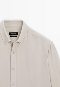 Camicia button-up beige chiaro in cotone. Presenta un colletto classico, una patta frontale con bottoni e maniche corte. Etichetta del marchio visibile.