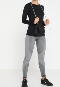 Top negro de manga larga, leggings grises texturizados con acentos de patrón y zapatillas deportivas negras; sosteniendo una cuerda para saltar.