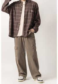 Camicia a scacchi marrone con taschino sul petto, abbinata a pantaloni cargo larghi color olivo con tasche laterali e sneakers con dettagli blu.