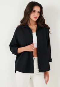 Camicia nera con bottoni, maniche lunghe, colletto e vestibilità comoda. Il tessuto appare liscio e leggero, abbinato a una maglietta bianca e pantaloni chiari.