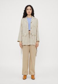 Gestreifte beige Jacke mit Fronttaschen und Knöpfen, getragen über einem hellblauen, verkürzten Pullover, kombiniert mit beigen Hosen und braunen Schuhen.