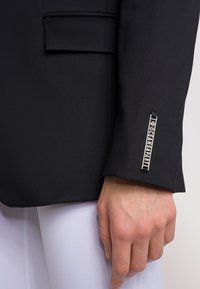 Czarna marynarka z wyprofilowanym kołnierzem i minimalnymi detalami kieszeni. Posiada srebrny akcent logo na mankiecie. Gładka tekstura i dopasowany krój.