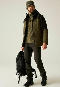 Hombre con chaqueta oliva y negra, suéter verde, pantalones oscuros y gorro, sosteniendo una mochila negra, de pie contra un fondo liso y claro.