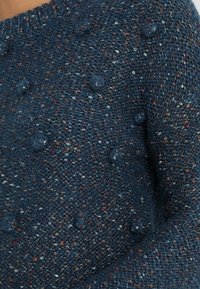 Pull-over tricoté bleu foncé avec des accents en relief à boucles et des nuances multicolores. Dispose d'un col rond et de poignets côtelés.