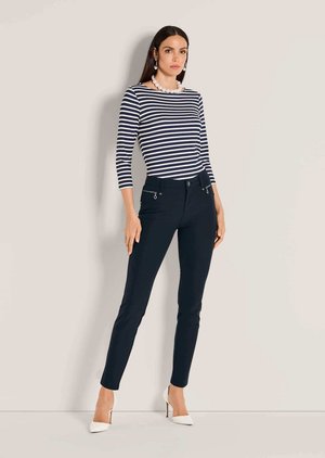 Vrouw die een marineblauw-wit gestreept shirt met lange mouwen draagt, marineblauwe slim fit broek met ritssluitingen in de zakken, witte hakken, parelketting en oorbellen.