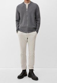 Giacca grigia con zip e colletto rotondo, abbinata a pantaloni beige chiaro e stivali neri con lacci. Il tessuto sembra morbido e strutturato.