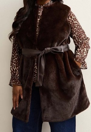 Woman wearing a sleeveless dark brown faux fur vest with a tied satin belt over a long-sleeve brown and white patterned blouse.