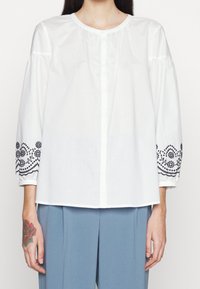 Blouse en coton blanc avec broderie florale noire sur des manches volumineuses, dotée d'un col rond et d'une fermeture à boutons à l'avant.