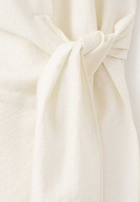 Tessuto color crema con una superficie morbida e strutturata. Presenta un dettaglio a nodo che crea un effetto arricciato. Nessun motivo o elemento di metallo visibile.