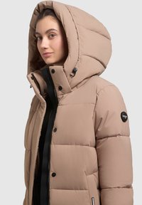 Beige puffer-takki, jossa on suuri huppu, vetoketjulla ja nepparilla suljettava, teksturoituja paneeleja ja tikkilogo vasemmassa hihassa.