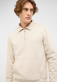 Beżowa bluza z półgolfem wykonana z miękkiego materiału, wyposażona w zamek na pół długości, kołnierz oraz przednią kieszeń z okrągłym logo.