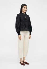 Γυναίκα που στέκεται φορώντας μαύρο μπλουζάκι με μακριά μανίκ ια και δαντέλα, εκρού παντελόνι cropped και σκούρα καφέ παπούτσια Mary Jane σε λευκό φόντο.