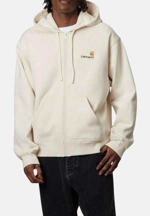 Hoodie zippé beige en tissu doux, avec une poche à l'avant, capuche avec cordon de serrage et un petit logo doré sur la poitrine gauche.