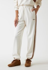 Witte joggingbroek met bruine zijkanten, voorzien van een elastische tailleband en een verstelbaar trekkoord. Inclusief merklogo op de heup.