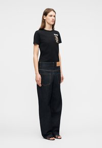 Mujer con camiseta negra con un gráfico de oso y jeans negros de pierna ancha con grandes bolsillos traseros y una etiqueta de cuero marrón claro.