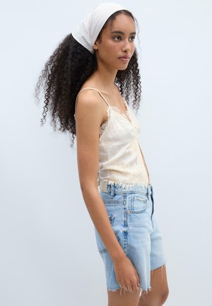 Jeune femme aux cheveux bouclés portant un foulard blanc, un caraco en dentelle et un short en jean bleu clair, tourné vers la droite sur un fond uni.