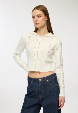Hoodie corto in maglia cable-knit bianca con zip frontale, cappuccio e motivi a rombi. Abbinato a jeans in denim blu scuro a vita alta.