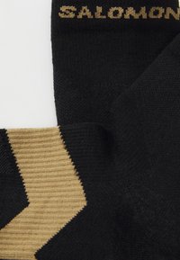 Calcetines negros con un acabado texturizado y un patrón en zigzag dorado. Nombre de la marca "SALOMON" tejido en dorado en la parte superior.