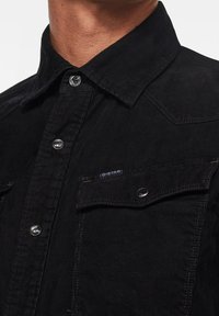 Czarna koszula z korduroyu z kołnierzykiem zapinanym na guziki, srebrnymi zatrzaskami i dwiema kieszeniami na piersiach. Charakteryzuje się teksturowanym materiałem oraz małym detalem z logo.