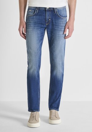 Uomo con jeans slim fit blu medio e sneaker beige, in piedi davanti a uno sfondo grigio chiaro uniforme.