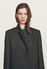 Czarny wełniany płaszcz o wysokim kołnierzu, z zapięciem na jeden guzik i dwoma przednimi kieszeniami; teksturowana tkanina z subtelnymi detalami wzoru.