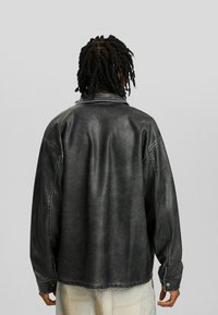 Černá džínová bunda s uvolněným střihem, viditelnými švy, límcem k sklopení a ozdobnými patentními knoflíky. Texturovaný, mírně opotřebovaný vzhled.