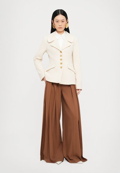 Blazer crema a medida con solapas prominentes y botones dorados, combinado con pantalones de pierna ancha marrones, que presentan una textura suave y un diseño estructurado.