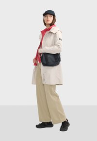 Veste imperméable beige clair avec des accents rouges, pantalons en khaki à coupe large et baskets noires. Un petit sac crossbody noir est porté.