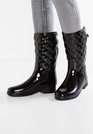 Bottes de pluie matelassées mi-mollet noires brillantes portées avec un jean slim gris clair sur fond blanc.