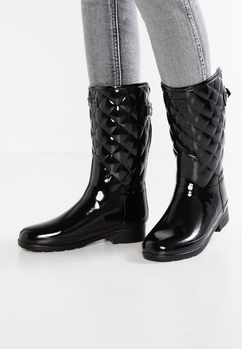 Bottes de pluie matelassées mi-mollet noires brillantes portées avec un jean slim gris clair sur fond blanc.