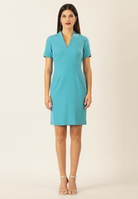 Türkises, kurzärmeliges Kleid mit V-Ausschnitt, strukturiertem Schnitt und knielangem Saum. Glattes Material, minimale Akzente, kombiniert mit silbernen Schuhen mit Knöchelriemen.