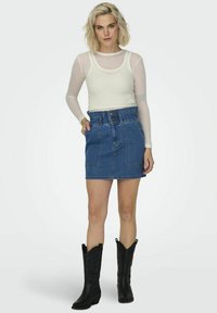 Fustă din denim cu talie elastică, două buzunare frontale și o nuanță clasică de albastru, combinată cu un top lung alb transparent cu mâneci lungi și cizme negre de cowboy.