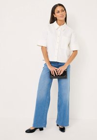 White short-sleeve shirt with a collar and pocket, paired with wide blue jeans and black glossy heels, holding a black clutch.