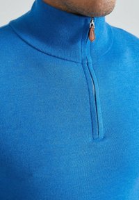 Pull en tricot bleu avec un col montant et une demi-fermeture éclair. Il possède une texture lisse et un tirant de fermeture éclair en cuir.