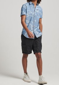 Hellblaue Kurzarmbluse mit weißem Vogelmuster, kombiniert mit schwarzen Cargo-Shorts und weißen Turnschuhen. Lässig, entspannte Passform.