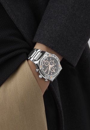 Ezüst színű fém krónográf óra texturált fekete számlappal, rózsaarany kiemelésekkel és nyolcszögletű tokkal. Barna szövetre van húzva a csuklón.